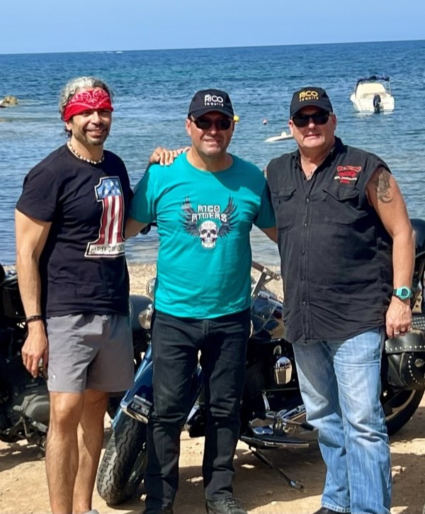 Rick with two friends beside their Harleys on a beach in Ibiza, the sea and anchored boats behind them